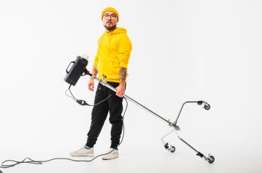 Sarı kapüşonlu ve bereli yaratıcı genç adam bir kamera tutuyor ve stüdyo ışıklandırma ekipmanları taşıyor. Modern fotoğrafçı sahne arkası konsepti