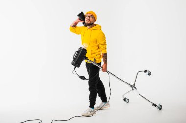 Sarı kapüşonlu ve bereli yaratıcı genç adam bir kamera tutuyor ve stüdyo ışıklandırma ekipmanları taşıyor. Modern fotoğrafçı sahne arkası konsepti