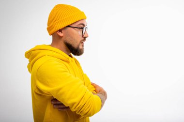 Sarı kapüşonlu ve bereli modern sakallı adamın beyaz arka planda duran kolları çaprazlanmış portresi. Moda ve kendine güvenen erkek tarzı.