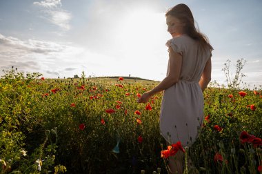 Gün batımında gelincik tarlasında yürüyen yaz elbisesi giymiş genç bir kadın. Doğal güzellik ve altın güneş ışığıyla huzurlu bir kırsal alan.