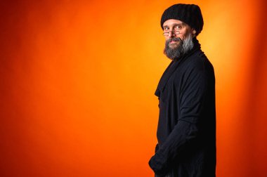 Gri sakallı, gözlüklü, gülümseyerek arkasını kollayan şık, yaratıcı, olgun bir adam. Canlı turuncu bir arkaplanda fotokopi alanı ile izole edilmiş.