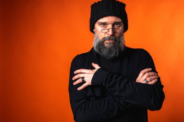 Sakallı, gözlüklü ve bereli, kollarını kavuşturmuş kendinden emin bir adam. Stüdyoda canlı turuncu arka planda sarsılmaz bir bakış.