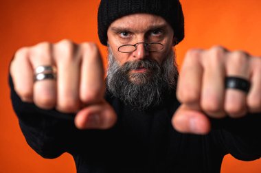 Sakallı, gözlüklü ve bereli kararlı bir adam. Kameraya doğru yumruk atıyor. Canlı turuncu üzerine yoğun stüdyo portresi, gücü ve mücadeleyi simgeliyor.