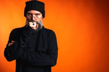 Sakallı, gözlüklü ve bereli etkileyici bir adam, göğüs çeneye. Canlı turuncu arka planda, stüdyo portresinin derinliklerinde.