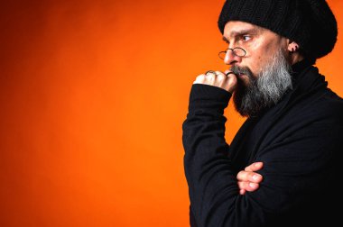 Uzun sakallı, gözlüklü ve bereli düşünceli bir adam. Canlı turuncu bir stüdyo portresi ile göğüs çeneye, derin düşünmeyi sembolize ediyor.