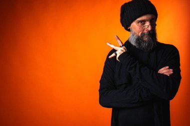 Sakallı, gözlüklü ve bereli, kollarını kavuşturmuş kendinden emin bir adam. Stüdyoda canlı turuncu arka planda sarsılmaz bir bakış.