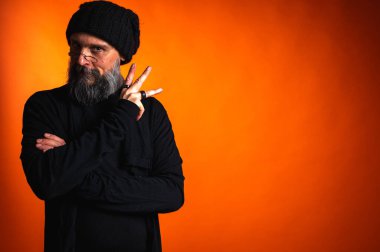 Sakallı, gözlüklü ve bereli, kollarını kavuşturmuş kendinden emin bir adam. Stüdyoda canlı turuncu arka planda sarsılmaz bir bakış.