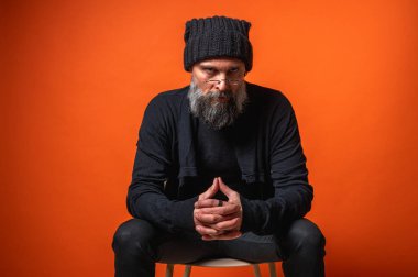 Siyah elbiseli ve bereli sakallı, parlak turuncu arka planda el ele oturan düşünceli, olgun bir adamın portresi. Şık erkek kavramı
