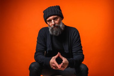 Siyah elbiseli ve bereli sakallı, parlak turuncu arka planda el ele oturan düşünceli, olgun bir adamın portresi. Şık erkek kavramı