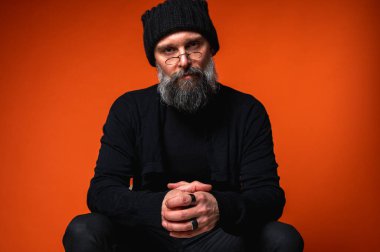 Siyah elbiseli ve bereli sakallı, parlak turuncu arka planda el ele oturan düşünceli, olgun bir adamın portresi. Şık erkek kavramı