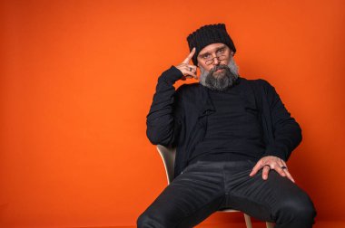 Sakallı, gözlüklü ve bereli etkileyici adam, göğüs göğüse. Canlı turuncu arka planda, stüdyo portresinin derinliklerinde.