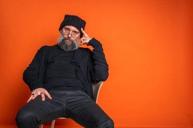 Sakallı, gözlüklü ve bereli etkileyici adam, göğüs göğüse. Canlı turuncu arka planda, stüdyo portresinin derinliklerinde.