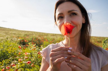 Gözleri kapalı, güneşli bir çayırda kırmızı bir gelincik çiçeğinin tadını çıkaran sakin bir kadın. Farkındalık ve doğa kavramı ile bağlantı