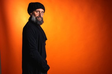 Gri sakallı, gözlüklü, gülümseyerek arkasını kollayan şık, yaratıcı, olgun bir adam. Canlı turuncu bir arkaplanda fotokopi alanı ile izole edilmiş.