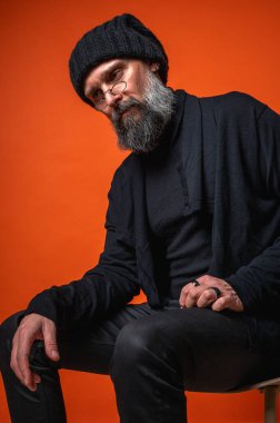 Gri sakallı ve gözlüklü, düşünceli, olgun bir adamın düşünceli portresi. Canlı turuncu bir arka planda izole edilmiş. Bilgelik, yaratıcılık ve uzmanlık kavramı