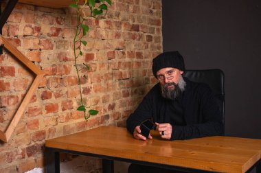 Kendine güvenen, sakallı modern bir iş adamı, çatı katında bir ofiste tuğla duvarlı bir masada oturuyor, katlanabilir bir akıllı telefon tutuyor. İş, teknoloji ve liderlik kavramı