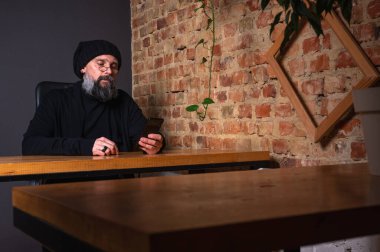 Kendine güvenen, sakallı modern bir iş adamı, çatı katında bir ofiste tuğla duvarlı bir masada oturuyor, katlanabilir bir akıllı telefon tutuyor. İş, teknoloji ve liderlik kavramı