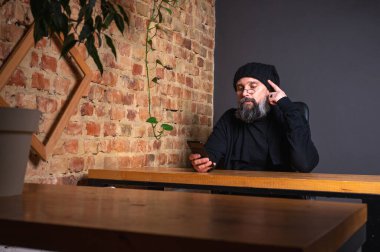 Kendine güvenen, sakallı modern bir iş adamı, çatı katında bir ofiste tuğla duvarlı bir masada oturuyor, katlanabilir bir akıllı telefon tutuyor. İş, teknoloji ve liderlik kavramı