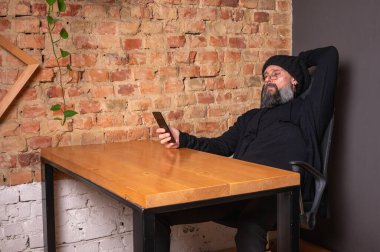 Kendine güvenen, sakallı modern bir iş adamı, çatı katında bir ofiste tuğla duvarlı bir masada oturuyor, katlanabilir bir akıllı telefon tutuyor. İş, teknoloji ve liderlik kavramı