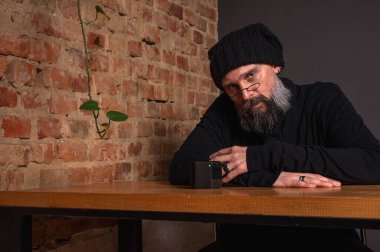 Ciddi sakallı, gözlüklü ve bereli bir hippinin portresi akıllı telefonuyla masada oturuyor. Çatı katında tuğla duvar arkasında yaratıcı bir serbest çalışan.