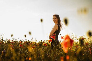Dalgalı saçlı güzel bir kadın gün batımında canlı bir haşhaş tarlasında duruyor, sıcak altın ışıkla yıkanmış, geriye dönüp izleyiciye bakıyor. Doğal güzellik ve neşe yakalar.