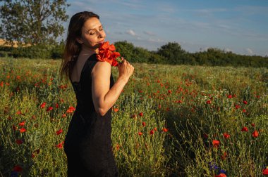 Siyah elbiseli bir kadın güneşli bir alanda canlı kırmızı haşhaş tutuyor, özgürlüğü, doğal güzelliği ve yaz neşesini somutlaştırıyor. Yaşam tarzı, moda ve duygusal temalar için ideal.