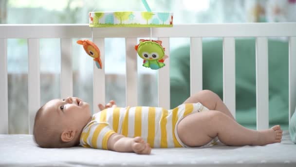 三个月大的婴儿在婴儿床里看着流动木马笑着 快乐的孩子新生儿在婴儿床里 儿童流动服务 — 图库视频影像
