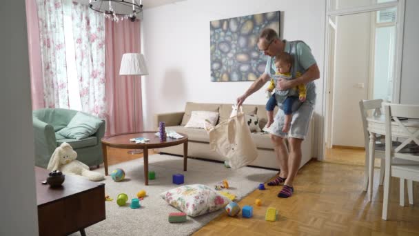 父はリビングルームで掃除をし 幼児の赤ちゃんと一緒にたくさんの散乱おもちゃを集めてスリングを持っています — ストック動画
