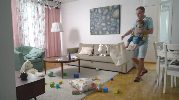父はリビングルームで掃除をし 幼児の赤ちゃんと一緒にたくさんの散乱おもちゃを集めてスリングを持っています — ストック動画