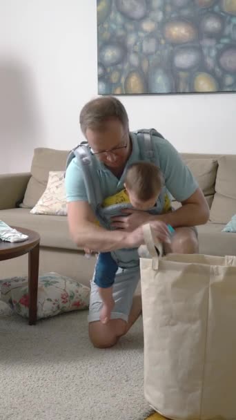 Apa Végzi Takarítást Nappaliban Összegyűjti Sok Szétszórt Játékot Csecsemőjével Parittyában — Stock videók