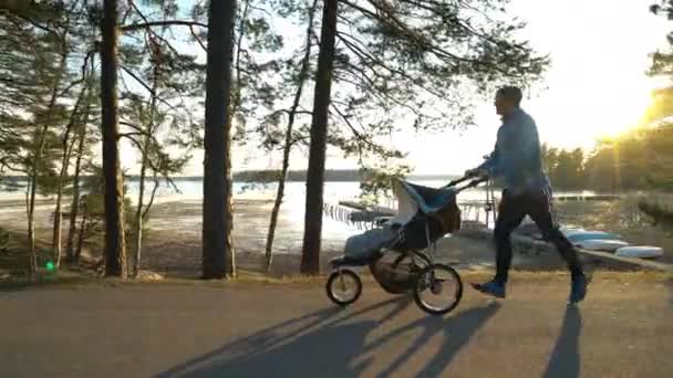 フィンランドの夕日の下で夜に堤防の公園の空の路地に沿って梅の赤ちゃんの馬車と男性ランナージョギング — ストック動画