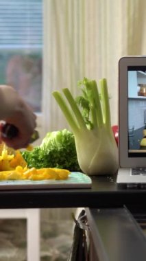 Lezzetli ve sağlıklı yiyeceklerle ilgili çevrimiçi video blogu öğreten genç bir kadın dizüstü bilgisayar kullanıyor ve ev mutfağında yemek pişiriyor..