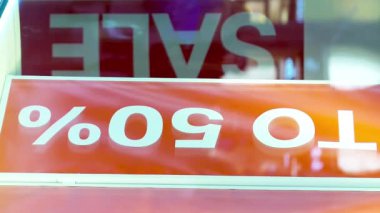 Şehrin merkez caddesindeki bir butik mağazasının vitrininde büyük bir indirimle ilgili afiş. Akşamın ilerleyen saatlerinde bir sürü tanınmayan yaya var.