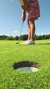 Golf oynayan olgun bir kadın. Golfçü golf topunu vuruyor ve golf deliğine vuruyor. Yeşil tepeleri ve mavi gökyüzü olan güneşli güzel bir manzara..