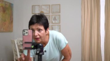 Olgun bir kadın evdeki sosyal medya hesabı için son moda dans hareketlerini kaydediyor. 50-60 yaşlarında bir kadın akıllı telefondan kısa video çekiyor. Sosyal medyayı, hikayeleri ve moda dansını kavrayın.