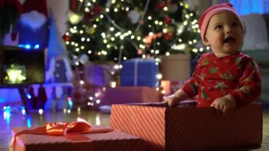 Noel arifesinde bir Noel ağacının arka planında kırmızı şapkalı, hediye kutusunda oturan mutlu çocuk. Mutlu tatiller, yeni yıl. Evde sıcak bir kış akşamı. Xmas zamanı.