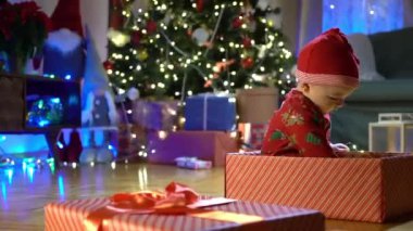 Noel arifesinde bir Noel ağacının arka planında kırmızı şapkalı, hediye kutusunda oturan mutlu çocuk. Mutlu tatiller, yeni yıl. Evde sıcak bir kış akşamı. Xmas zamanı.