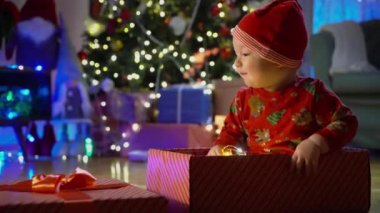 Noel arifesinde bir Noel ağacının arka planında kırmızı şapkalı, hediye kutusunda oturan mutlu çocuk. Mutlu tatiller, yeni yıl. Evde sıcak bir kış akşamı. Xmas zamanı.