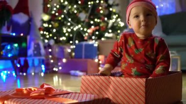 Noel arifesinde bir Noel ağacının arka planında kırmızı şapkalı, hediye kutusunda oturan mutlu çocuk. Mutlu tatiller, yeni yıl. Evde sıcak bir kış akşamı. Xmas zamanı.