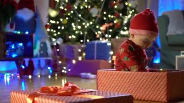Noel arifesinde bir Noel ağacının arka planında kırmızı şapkalı, hediye kutusunda oturan mutlu çocuk. Mutlu tatiller, yeni yıl. Evde sıcak bir kış akşamı. Xmas zamanı.