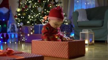 Noel arifesinde bir Noel ağacının arka planında kırmızı şapkalı, hediye kutusunda oturan mutlu çocuk. Mutlu tatiller, yeni yıl. Evde sıcak bir kış akşamı. Xmas zamanı.