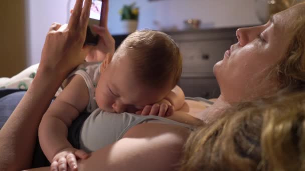 スマートフォンを使用しながら ベッドルームで母親の胸に寝ている赤ちゃんの息子 — ストック動画