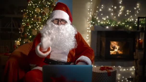 圣诞老公公在圣诞前夕坐在家中的笔记本电脑上 用虚拟视频在线聊天 把礼物握在手里 祝孩子们和成年人圣诞快乐和新年快乐 — 图库视频影像