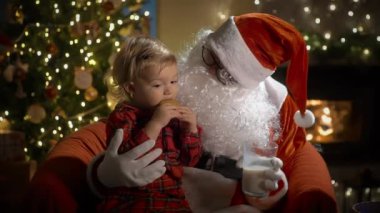Pijamalı ve Noel Baba 'lı küçük bir çocuk Noel kurabiyelerini sütle yer ve Noel hediyelerini Noel ağacı ve şöminesi olan süslü bir oturma odasında yerleştirir.