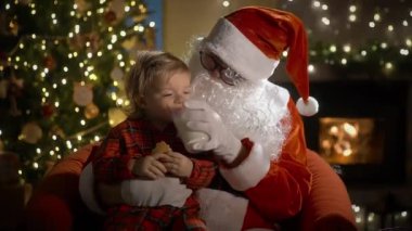 Pijamalı ve Noel Baba 'lı küçük bir çocuk Noel kurabiyelerini sütle yer ve Noel hediyelerini Noel ağacı ve şöminesi olan süslü bir oturma odasında yerleştirir.