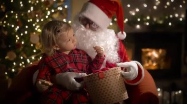 Pijamalı ve Noel Baba 'lı küçük bir çocuk Noel kurabiyelerini sütle yer ve Noel hediyelerini Noel ağacı ve şöminesi olan süslü bir oturma odasında yerleştirir.