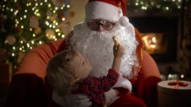 Pijamalı ve Noel Baba 'lı küçük bir çocuk Noel kurabiyelerini sütle yer ve Noel hediyelerini Noel ağacı ve şöminesi olan süslü bir oturma odasında yerleştirir.