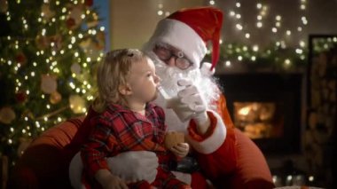 Pijamalı ve Noel Baba 'lı küçük bir çocuk Noel kurabiyelerini sütle yer ve Noel hediyelerini Noel ağacı ve şöminesi olan süslü bir oturma odasında yerleştirir.