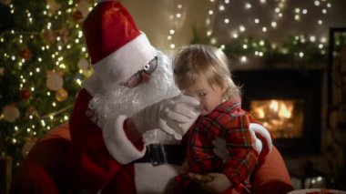 Pijamalı ve Noel Baba 'lı küçük bir çocuk Noel kurabiyelerini sütle yer ve Noel hediyelerini Noel ağacı ve şöminesi olan süslü bir oturma odasında yerleştirir.