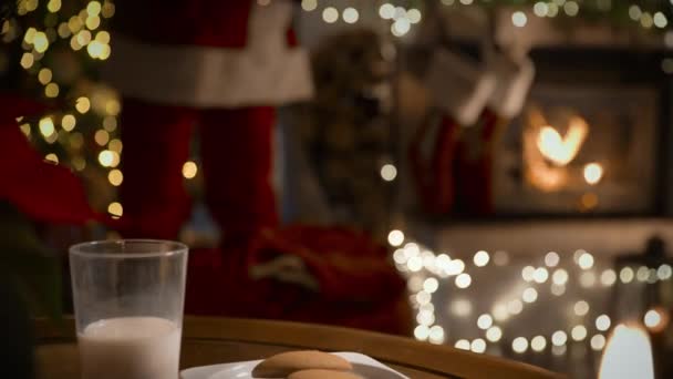 サンタクロースは暖炉の上に靴下にクリスマスプレゼントを置き ジンジャークッキーを食べ クリスマスツリーで装飾されたリビングルームでミルクを飲む — ストック動画